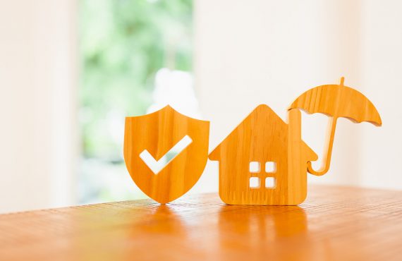 Hausminiatur aus Holz steht auf einem Tisch, wird durch einen Regenschirm geschützt. Daneben steht ein Wappen mit Haken.