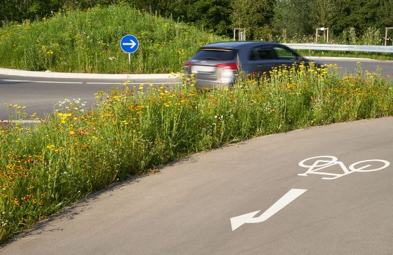 Ein graues Auto fährt durch einen Kreisverkehr. Im Vordergrund sieht man einen abbiegenden Radweg.