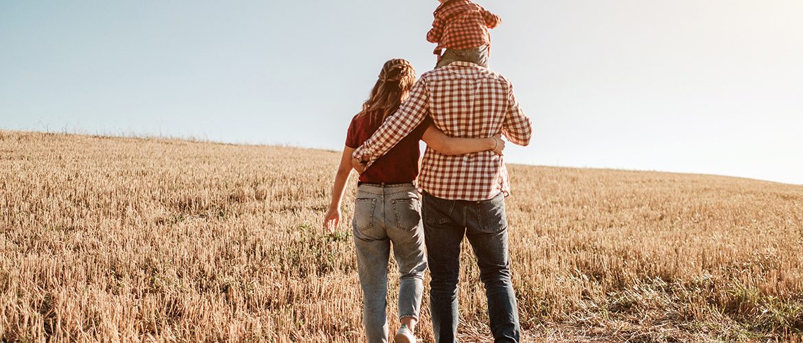 Eine junge Familie steht mit dem Rücken zugewandt auf einem abgeernteten Getreidefeld und schaut positiv in die Zukunft.
