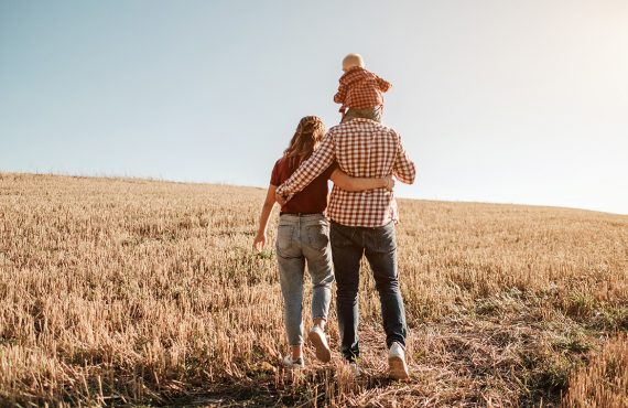 Eine junge Familie steht mit dem Rücken zugewandt auf einem abgeernteten Getreidefeld und schaut positiv in die Zukunft.