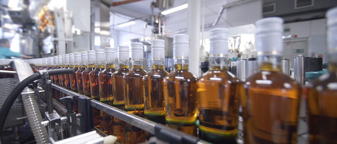 Auf einer industriellen Abfüllanlage stehen Flaschen in einer Reihe. Der Inhalt ist eine leicht braune Flüssigkeit.