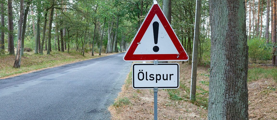Landstraße im Wald. Ein Verkehrsschild steht am rechten Rand und weißt auf eine Ölspur hin.