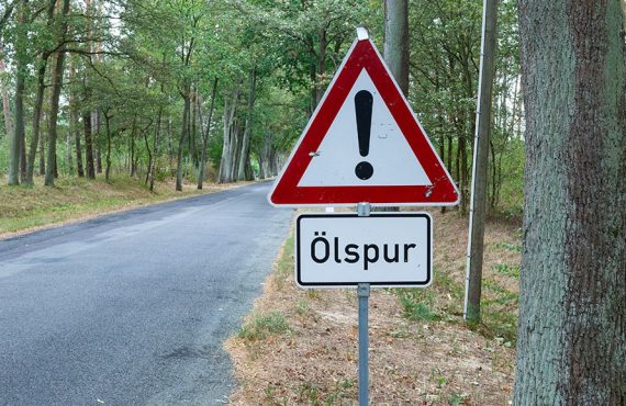 Landstraße im Wald. Ein Verkehrsschild steht am rechten Rand und weißt auf eine Ölspur hin.